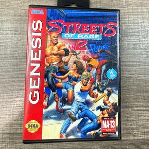 May include: A SEGA Genesis video game cartridge for Streets of Rage 2. The cover art features a group of characters in action, with the game's title in a stylized font. The cartridge is in a black case with a red spine.