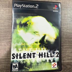 May include: A black PlayStation 2 game case featuring the game "Silent Hill 2." The cover art depicts a distorted face in shades of green and white. The game case has the PlayStation 2 logo and the ESRB rating.