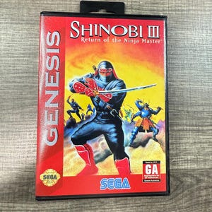 May include: A red and black SEGA Genesis game cartridge for Shinobi III: Return of the Ninja Master. The cover art features a ninja in a black suit with a red mask and boots, holding a sword. The SEGA logo is at the bottom.