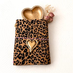 Può includere: Una copertina per taccuino rettangolare con motivo leopardato marrone e nero. Un sottile elastico rosa avvolge la copertina, fissando un ciondolo dorato a forma di cuore. Dietro il taccuino si trovano un piattino dorato a cuore e fiori secchi.