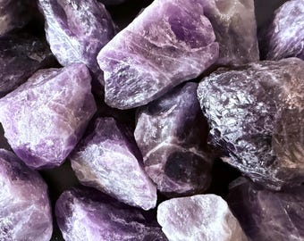 May include: A close-up shot of several rough-cut amethyst crystals. The crystals vary in size and shape, with a range of purple hues from light lavender to deep violet. The raw stones have a natural, textured appearance.