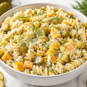 May include: A white bowl filled with rotini pasta salad tossed in a creamy white dressing. The dish contains diced orange cheddar cheese cubes and sliced green pickles, garnished with fresh green dill sprigs throughout the mixture.