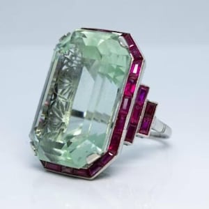 May include: A large, rectangular green gemstone ring with a silver band. The ring is accented with small, rectangular red gemstones around the green stone and on the side of the band. The ring is set against a white background.