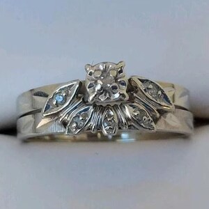 May include: A silver engagement ring set with a square-cut diamond center stone and smaller accent diamonds. The band features a floral design with leaf-shaped details. The ring set is displayed in a white ring box.