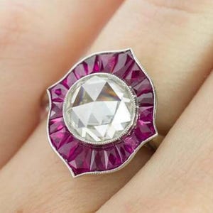 Peut inclure: Une bague de style vintage avec une grosse pierre centrale claire et facettée, entourée d'un halo de petites pierres précieuses rouge foncé. La bague a un anneau argenté et un bord décoratif festonné.