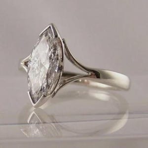 May include: A silver-toned ring featuring a large, marquise-cut clear gemstone set in a prong setting. The band has a split-shank design that tapers as it connects to the central stone, creating a sleek and polished appearance with a reflective finish.