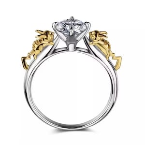 May include: A silver ring with a large, clear gemstone. The ring features two gold-colored rabbit figures on either side of the gemstone. The ring is set against a white background.