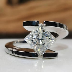 May include: A contemporary silver ring with a large, square-cut diamond as its centrepiece. The diamond is set in an open, modern design. The band has a geometric shape, enhancing the ring's contemporary style. The ring is displayed on a white surface.