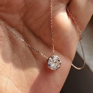 Puede incluir: Collar de oro rosa con un colgante de diamante redondo con talla brillante. El diamante está engastado en un bisel y cuelga de una delicada cadena. El collar se muestra sobre un fondo suave.