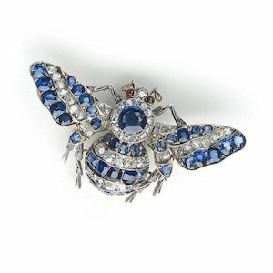 May include: A jeweled bee brooch with blue and white gemstones. The wings are adorned with blue stones, while the body features alternating bands of blue and white gems. Small red stones accent the head.