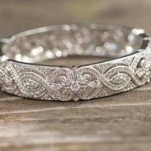 Puede incluir: Un brazalete de plata con bisagras con intrincados filigranas y brillantes piedras preciosas transparentes. El brazalete presenta un diseño floral y de desplazamiento detallado, sobre un fondo texturizado. La joya se exhibe sobre una superficie de madera.
