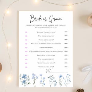 以下が含まれることがあります： エレガントな筆記体で「Bride or Groom」と書かれた白いカード。カードには、さまざまなことで誰が優れているかを推測するための質問があり、回答を丸で囲むスペースがあります。青と薄紫の花柄のアクセントが下部を飾っています。