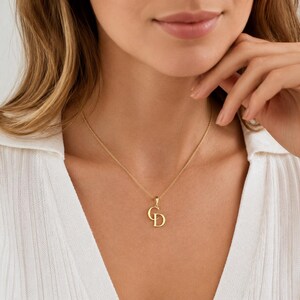 May include: A gold-toned necklace featuring a pendant with the letters C and D intertwined. The pendant hangs from a delicate gold-toned chain that rests against the collarbone. The letters are rendered in a smooth, polished metal finish.