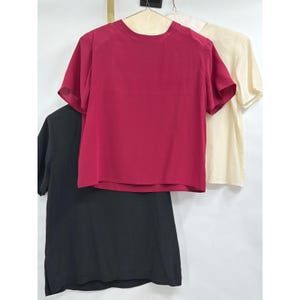 May include: A short-sleeved crew neck top in a vibrant magenta color, displayed alongside a black short-sleeved top and a cream-colored short-sleeved top. Each garment features a simple, solid-colored design with a rounded neckline and straight hem.