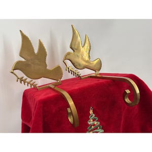 Könnte beinhalten: Ein Paar Strumpfhalter aus poliertem Messing, die jeweils eine fliegende Taube mit einem Olivenzweig darstellen. Die Metallfiguren sind an gebogenen, beschwerten Haken befestigt, die zum Aufhängen an einer Kaminsims- oder Regaloberfläche dienen.