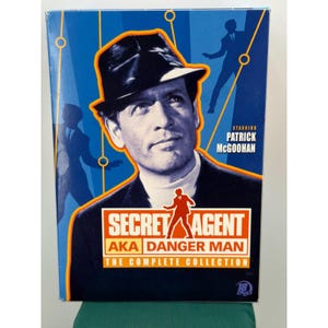 May include: A rectangular DVD box set titled Secret Agent AKA Danger Man The Complete Collection. The cover features a black and white portrait of Patrick McGoohan wearing a hat, set against a blue background with orange lines and silhouettes.