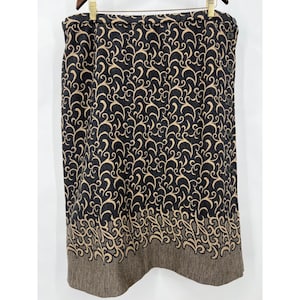 Könnte beinhalten: Ein knielanger Rock mit schwarzem Hintergrund und einem beigefarbenen Wirbelmuster. Der Rock hat einen braunen Saum und einen Knopfverschluss an der Seite. Der Rock hängt an einem Holzbügel.