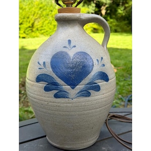 May include: A light gray stoneware jug lamp featuring a hand-painted blue heart motif with decorative leaf accents. The jug has a single side handle, a narrow neck with a wooden stopper, and a brown electrical cord extending from the base.