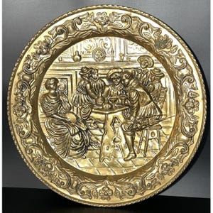 May include: A round brass decorative plate featuring a high-relief embossed scene of figures gathered around a table in an interior setting. The wide border is decorated with intricate floral and scroll patterns, and the metal has a polished finish.