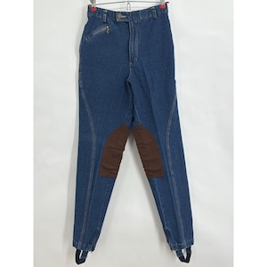 May include: Blue denim riding pants with brown suede knee patches. The pants have a high waist, a zipper pocket, and stirrups at the ankles. The pants are hanging on a hanger against a white background.