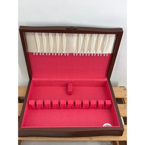 Pode incluir: Um estojo de armazenamento retangular de madeira com acabamento exterior castanho escuro. O interior é forrado com tecido rosa brilhante, apresentando uma fila de doze ranhuras individuais para talheres e um painel de tecido plissado creme.