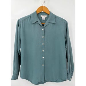May include: A teal button-down shirt with long sleeves, hanging on a wooden hanger. The shirt has a classic collar and white buttons down the front. The tag reads "Talbots Petite".