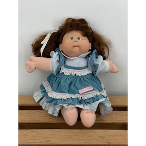Op de afbeelding: Een vintage Cabbage Patch Kids pop met bruin krullend haar en blauwe ogen. De pop draagt een blauwe jurk met witte kanten rand en een witte strik. De naam van de pop is zichtbaar op de jurk.