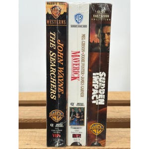 Pode incluir: Três fitas VHS: "The Searchers" com John Wayne, "Maverick" com Mel Gibson e Jodie Foster, e "Sudden Impact" da Coleção Clint Eastwood. As fitas estão em embalagem plástica e apresentam o logotipo da Warner Bros.
