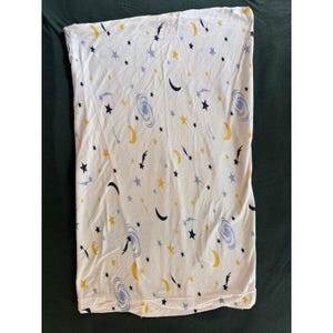 Può includere: Un telo in tessuto rettangolare bianco con un motivo celeste ripetitivo composto da lune crescenti gialle, stelle blu scuro, stelle azzurre e galassie a spirale azzurre sparse casualmente su tutta la superficie.