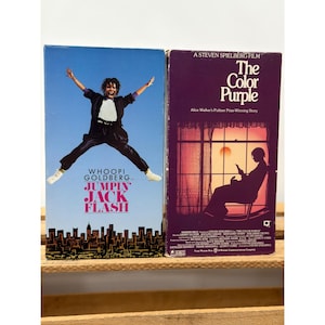 Może przedstawiać: Dwie kasety VHS. Jedna przedstawia Whoopi Goldberg w filmie "Jumpin' Jack Flash" z niebieskim tłem nieba. Druga to "Kolor purpury" z fioletową okładką i sylwetką osoby czytającej.