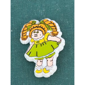 May include: A white plastic pin featuring a cartoon doll with orange hair in two braids, wearing a bright yellow dress with a green skirt overlay, yellow shoes, and white socks with ruffles. The doll has green eyes and a small green flower detail.
