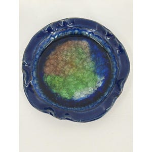 Puede incluir: Un plato redondo de cerámica con un borde azul profundo, ondulado y elevado. El centro presenta un esmalte cristalino texturizado con parches mezclados de verde, marrón y blanco, creando una apariencia multicolor y agrietada dentro del borde azul.