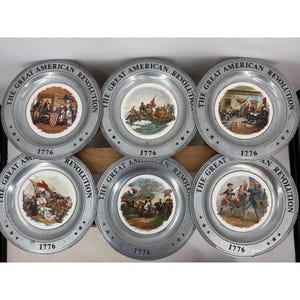 May include: A set of six circular metal plates featuring historical scenes from the American Revolution. Each plate has a silver-toned rim with the text THE GREAT AMERICAN REVOLUTION and 1776 printed on it, surrounding a central colour illustration.