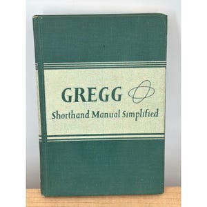Può includere: Un libro vintage con copertina rigida intitolato "GREGG Shorthand Manual Simplified". Il libro ha una copertina verde scuro con un pannello rettangolare verde chiaro al centro. Il titolo e un simbolo stilizzato sono stampati in nero. Il dorso del libro è visibile.