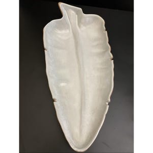 May include: A ceramic serving tray shaped like a large leaf with a glossy off-white finish. The dish features a central vein running the length of the piece, with subtle indentations along the edges that mimic the natural structure of a leaf.