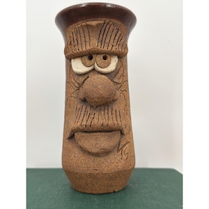 May include: A brown ceramic vase with a textured, gritty surface featuring a sculpted face with prominent eyebrows, round eyes, a large nose, a mustache, and a mouth. The rim is smooth and dark brown, and the initials KG are etched on the side.