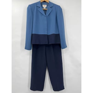 May include: A two-piece women's suit in shades of blue. The jacket is two-toned, with a lighter blue upper section and a darker blue lower section. The matching pants are a solid, dark blue color. The suit is displayed on a wooden hanger.