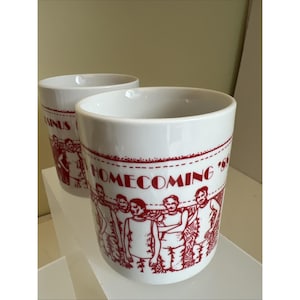 May include: Two white ceramic mugs with a red design. The mugs feature the text "HOMECOMING '89" and a graphic of people. The mugs are cylindrical with a smooth, glossy finish.