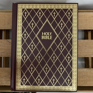 Könnte beinhalten: Eine burgunderfarbene Bibel mit goldgeprägten Kreuzen und Rautenmustern. Der Titel "HOLY BIBLE" ist mittig auf dem Einband platziert. Die Kanten sind goldfarben eingefasst. Das Buch ist geschlossen und liegt auf einer Holzoberfläche.