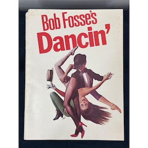 以下が含まれることがあります： ボブ・フォッシーの「ダンシン」のヴィンテージプログラム表紙。上部に太い赤文字でタイトルが記されている。黒い衣装を身にまとい、山高帽を被ったダンサーと赤いハイヒールを履いたダンサーが、クリーム色の背景に対してダイナミックなポーズをとっているイラストが描かれている。