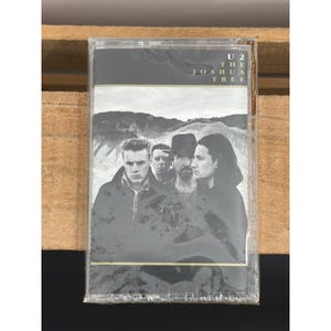 May include: A U2 The Joshua Tree cassette tape in a clear plastic case. The cover features a black and white photograph of the band members standing in a desert landscape. The text U2 THE JOSHUA TREE is printed in white in the upper right corner.