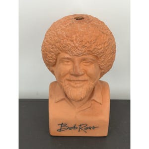 May include: A terracotta-colored ceramic planter shaped like the head and shoulders of Bob Ross, featuring his signature curly hair and beard. The base of the bust displays the name Bob Ross in black script lettering, with an opening at the top.
