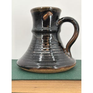 May include: A ceramic mug with a wide, flared base and a narrow neck, featuring a glossy dark brown and black glaze with visible horizontal ridges along the body. The mug has a single curved handle on the right side and a raw, unglazed tan base.