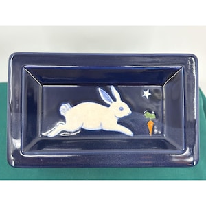 May include: A rectangular ceramic tray with a glossy dark blue finish. The center features a raised, light-colored rabbit in a running pose, a small white star, and a single orange carrot with a green top, all set against the deep blue background.