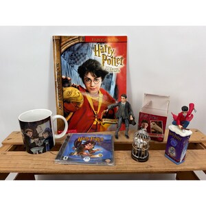 May include: A collection of Harry Potter merchandise including a colouring book, a white ceramic mug with character illustrations, a video game case, a posable action figure, a small birdcage ornament, and a decorative figurine on a base.