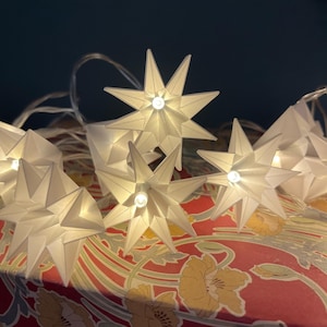 Può includere: Una stringa di luci bianche a forma di stella origami. Ogni stella ha una piccola luce brillante al centro. Le luci sono appoggiate su una superficie rossa con un motivo floreale, creando un bagliore caldo. Lo sfondo è di colore verde acqua scuro.