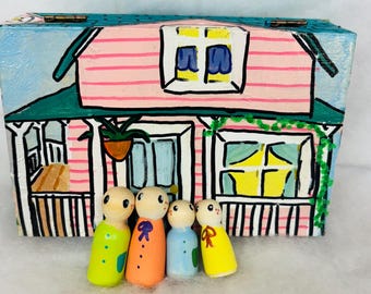 Puede incluir: Una caja de madera pintada a mano que representa una casa rosa con un tejado verde y un cielo azul. Cuatro muñecos de madera están de pie delante, vestidos de amarillo, naranja, azul y verde. La casa tiene un porche y una planta colgante.