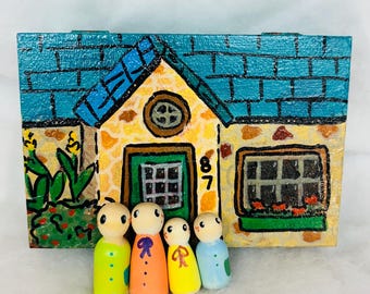 Puede incluir: Una casa de madera pintada a mano con un tejado de tejas azules, una fachada de piedra amarilla y el número 87 sobre la puerta verde. Cuatro muñecos de madera de colores brillantes están delante de la casa.