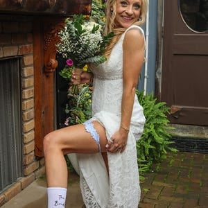 Könnte beinhalten: Eine Braut in einem weißen Spitzen-Hochzeitskleid, die einen Strauß weißer Blumen und Grünzeug hält. Sie trägt eine hellblaue Strumpfband, weiße Socken mit dem Aufdruck "The Bride" und weiße Turnschuhe mit grünen Schnürsenkeln und Perlen.