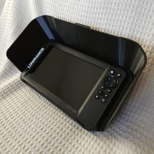 May include: A black Lowrance Eagle 3D fish finder unit with a protective black visor. The unit has a dark screen, control buttons, and the Lowrance brand name is visible on the visor. The fish finder is designed for marine use.